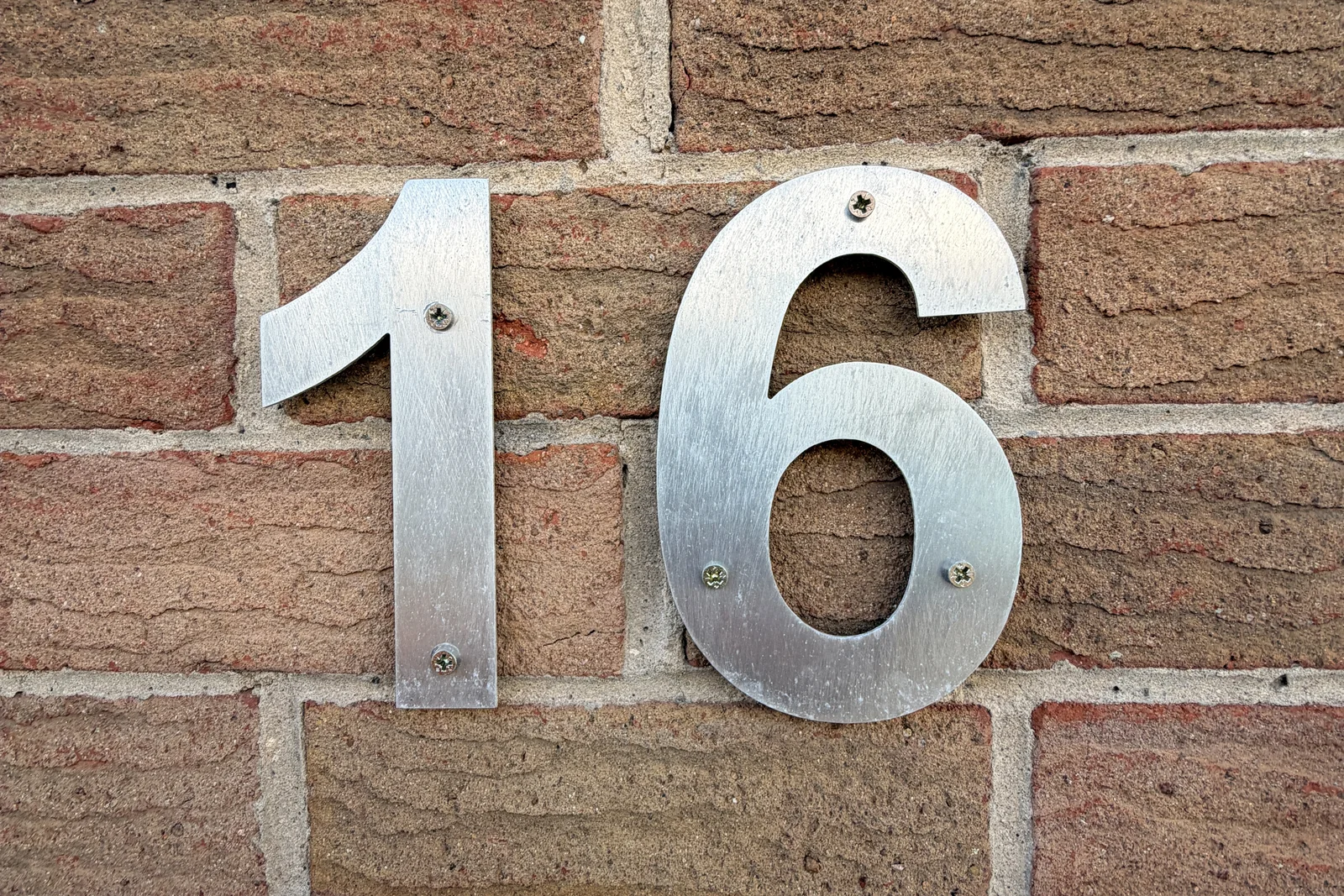 Brushed aluminium cut-out house number 16 fixed to a brick wall